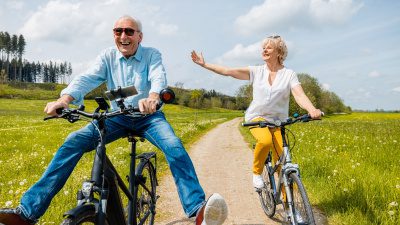 Två seniorer cyklar leende på en sommarväg